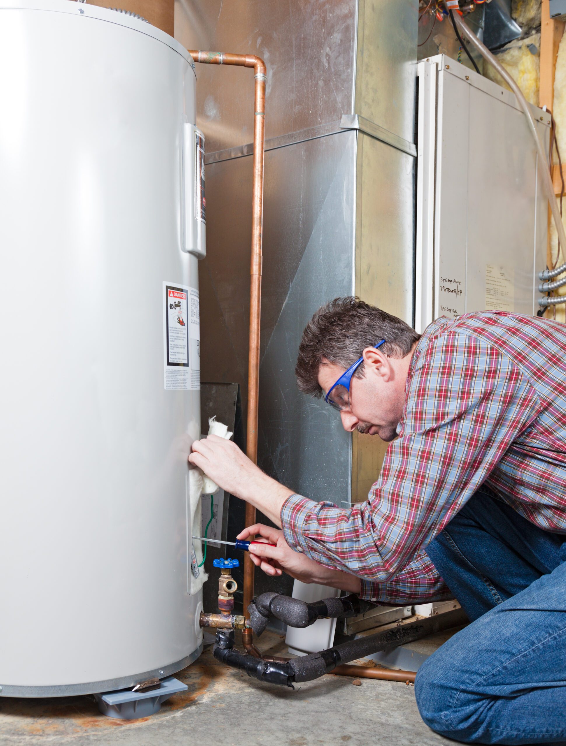 A man wearing safety glasses kneels to adjust a valve on a water heater in a utility room., Experienced plumber testing water pressure at a Cantonment - FL home with diagnostic tools, Plumber inspecting drain connections under a Milton - FL kitchen sink with a flashlight