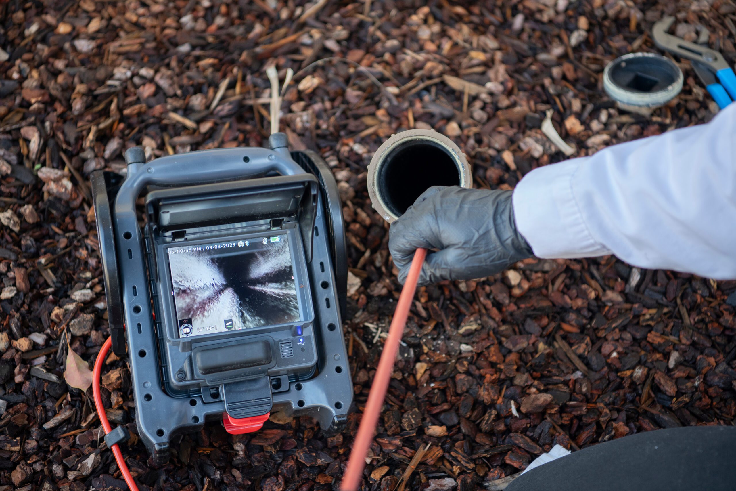 A person uses a camera inspection device to examine the inside of an outdoor pipe surrounded by bark mulch., Skilled plumber tracing an active leak behind a wall in a Pensacola - FL residence