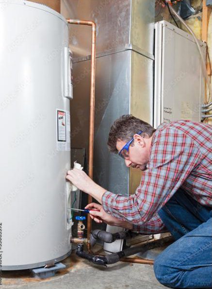 Man in glasses and plaid shirt inspects or repairs a water heater in a basement, using tools near copper pipes and furnace equipment., Plumber diagnosing drain and supply issues inside a Fort Walton Beach - FL utility room