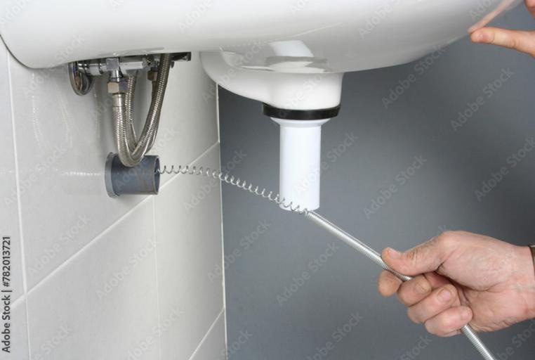A person uses a plumbing snake tool to unclog a sink drain pipe., Experienced plumber diagnosing a pipe leak inside an Escambia County - FL utility room