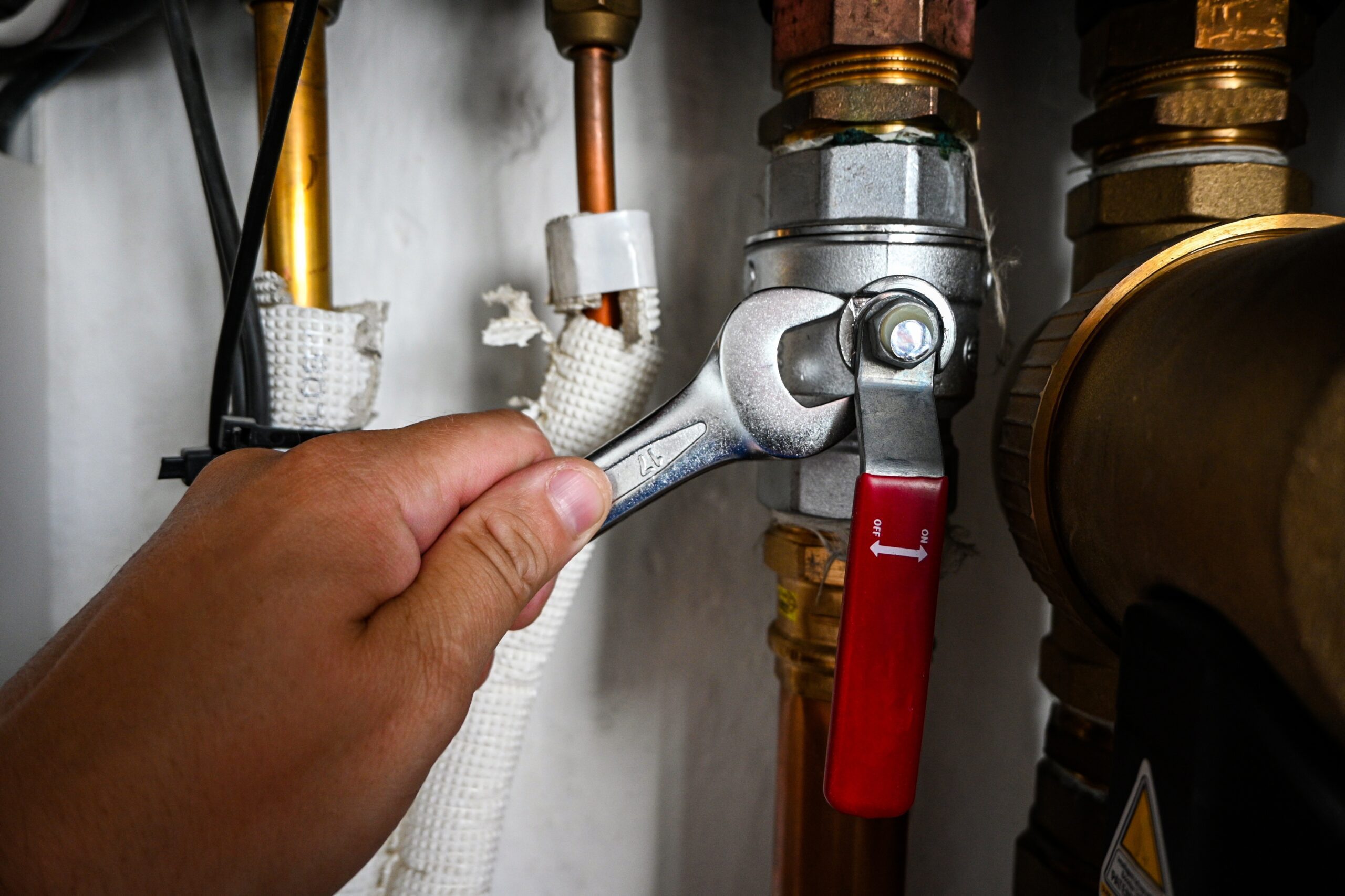 A hand uses a wrench to tighten or loosen a metal valve connected to copper pipes, with a red-handled lever visible., Plumber in Santa Rosa County - FL replacing a bathroom shutoff valve during a residential repair