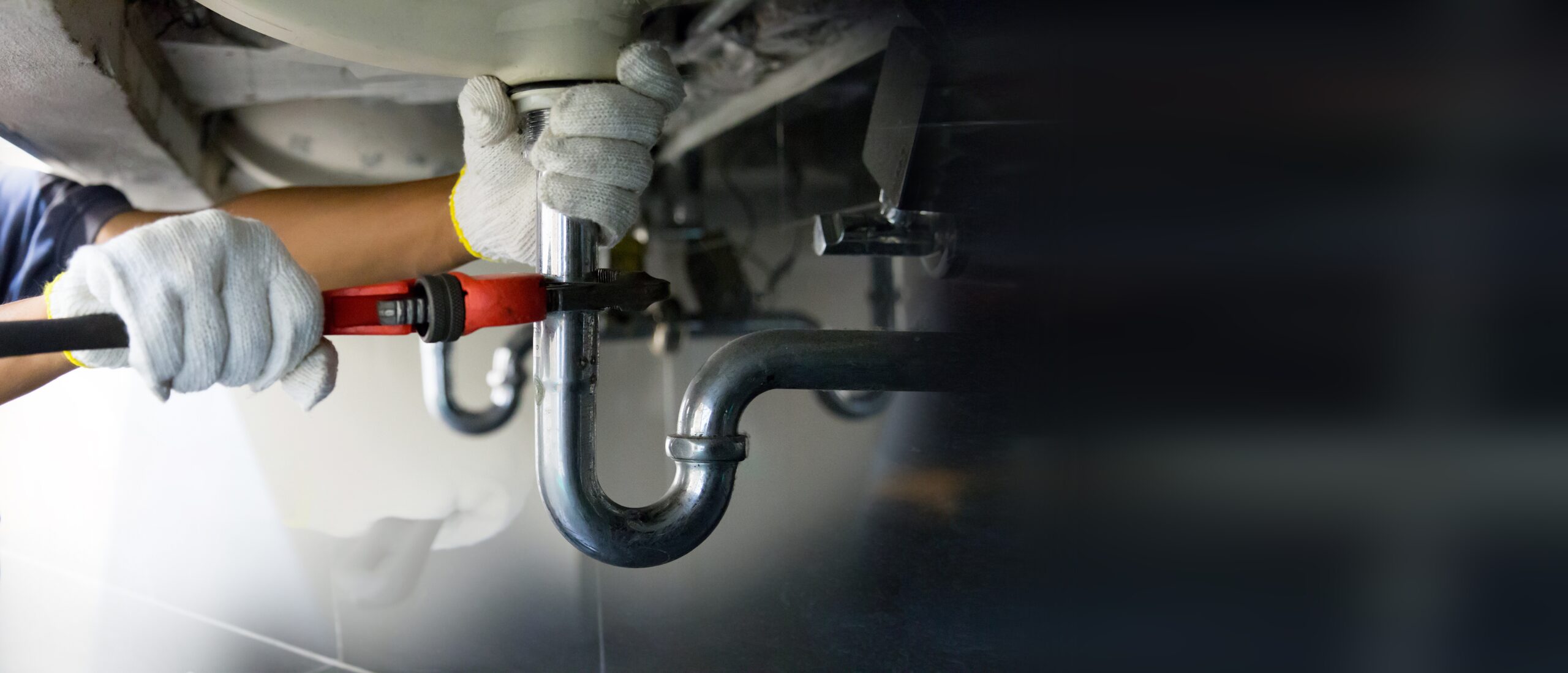 A person wearing gloves uses a wrench to tighten the pipe under a sink, performing plumbing maintenance., Plumber tracing a water line issue inside a Century - FL utility room with hand tools